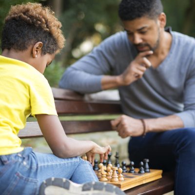 Father and daughter playing chess on the bench in city park.